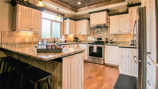 A kitchen with white cabinets, granite countertops, a central island with bar stools, stainless steel appliances, and a wooden ceiling. - Luxury Cabin,Golf Course,Yellowstone, Stunning Mountain Views,Tubing, Rafting, (Island Park)