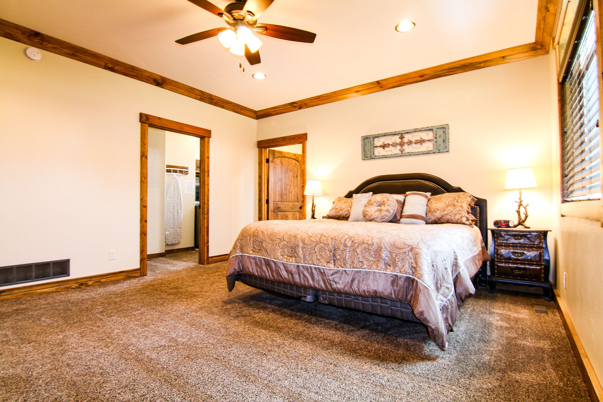 A bedroom with a large bed, wooden ceiling beams, a ceiling fan, and a nightstand with a lamp.