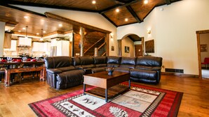 A spacious living room with a large leather sectional sofa, a wooden coffee table, and a red and white patterned rug. A kitchen with wooden cabinets and a dining area with a wooden table and chairs are visible in the background. - Luxury Cabin,Golf Course,Yellowstone, Stunning Mountain Views,Tubing, Rafting, (Island Park)