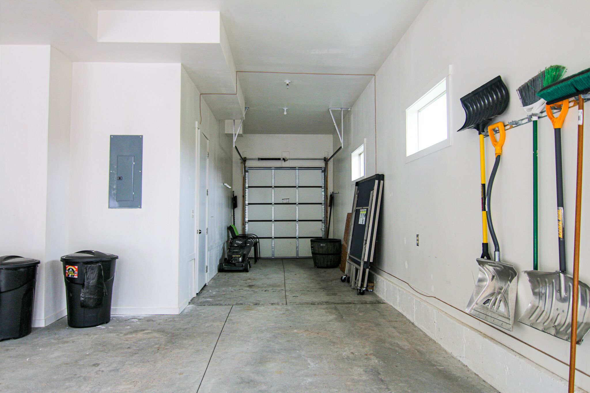 Eine Garage mit Betonboden, geschlossenem Garagentor und verschiedenen Reinigungsmitteln wie Besen und Mopps an der Wand.