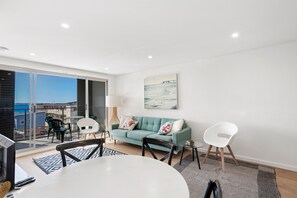 A modern living room with a teal sofa, a white dining table, and a glass coffee table.