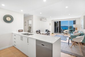 A modern kitchen with a dishwasher, a dining area with a table and chairs, and a balcony with a view of the sea. - The Shoal Apartment 310 Coastal Escape with Views (Shoal Bay)