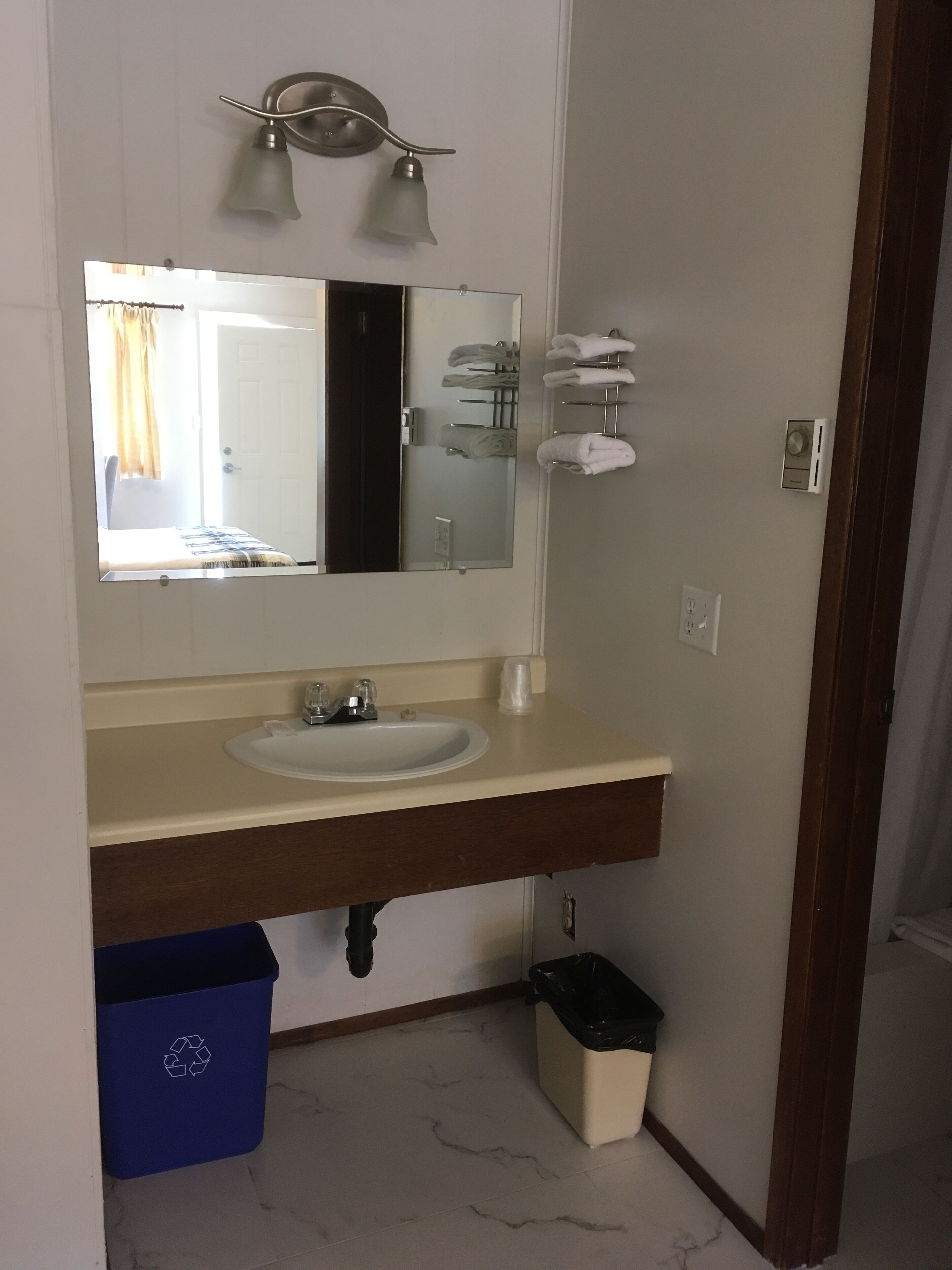 Un baño con un lavamanos blanco, un espejo y un perchero. Hay un cubo de basura azul y uno negro debajo del lavamanos.