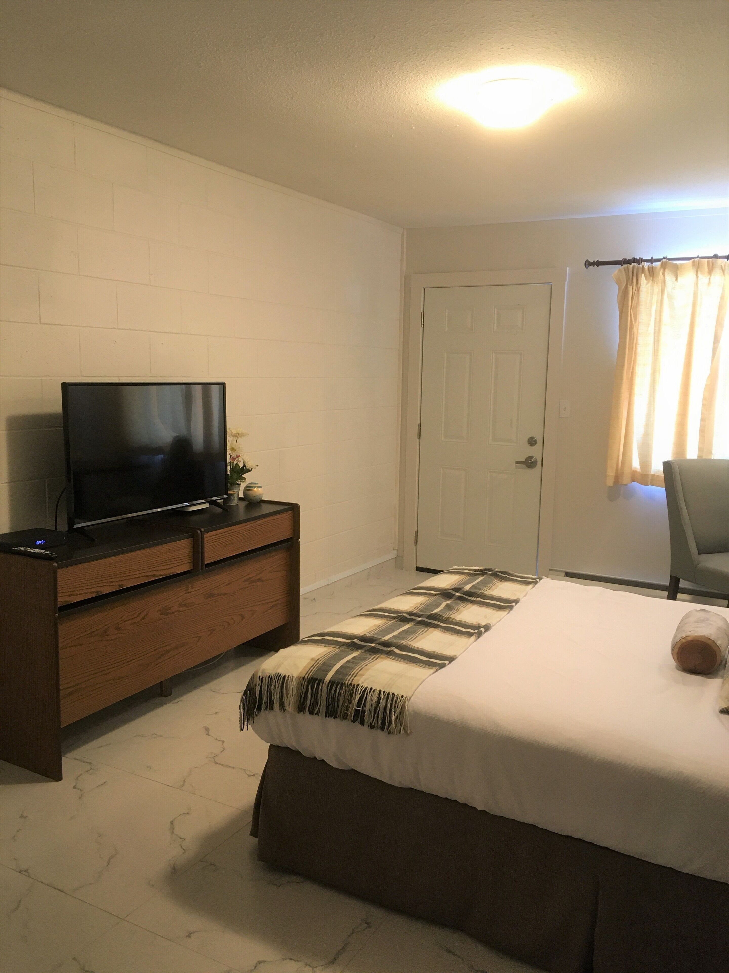 Un dormitorio con cama, televisor sobre una cómoda de madera, una puerta y una ventana con cortinas.