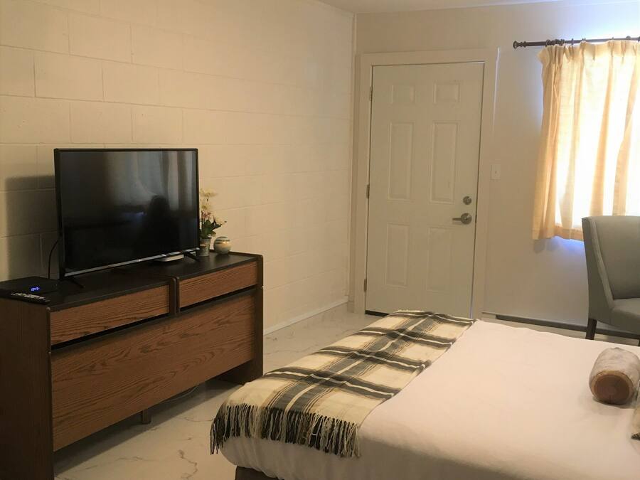 A bedroom with a bed, a TV on a wooden dresser, a door, and a window with curtains.