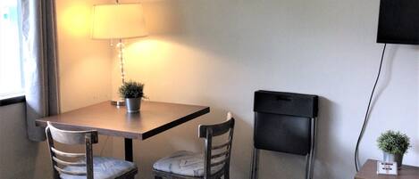 Habitación de hotel con cama, una mesa con dos sillas, lámpara, televisor y una planta pequeña en una mesita.