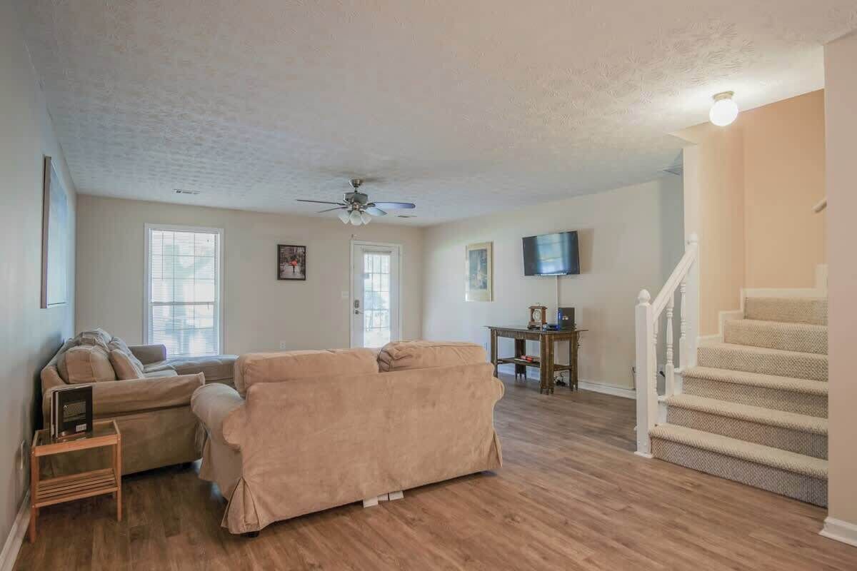 A living room with a beige sofa, a ceiling fan, a flat-screen TV, and a staircase.