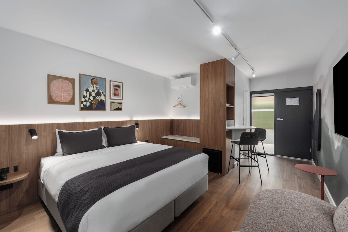 A modern hotel room with a large bed, bedside tables, a desk, and a chair. There are wall-mounted lights, a coat rack, and framed artwork.
