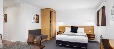 A hotel room with a bed, a desk, a chair, a wardrobe, and a small table.