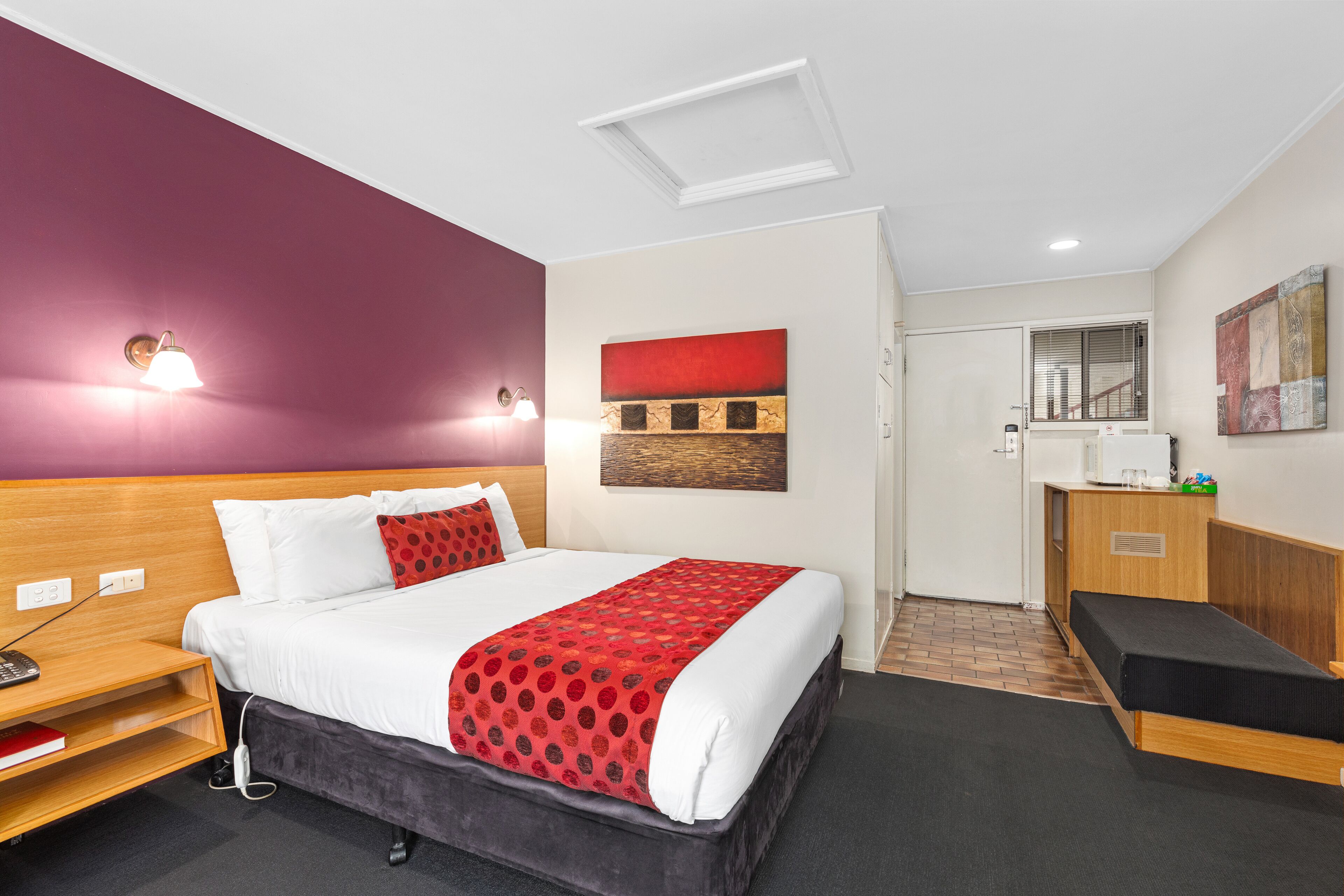 A hotel room with a bed, bedside table, lamp, artwork, and a desk with a chair.