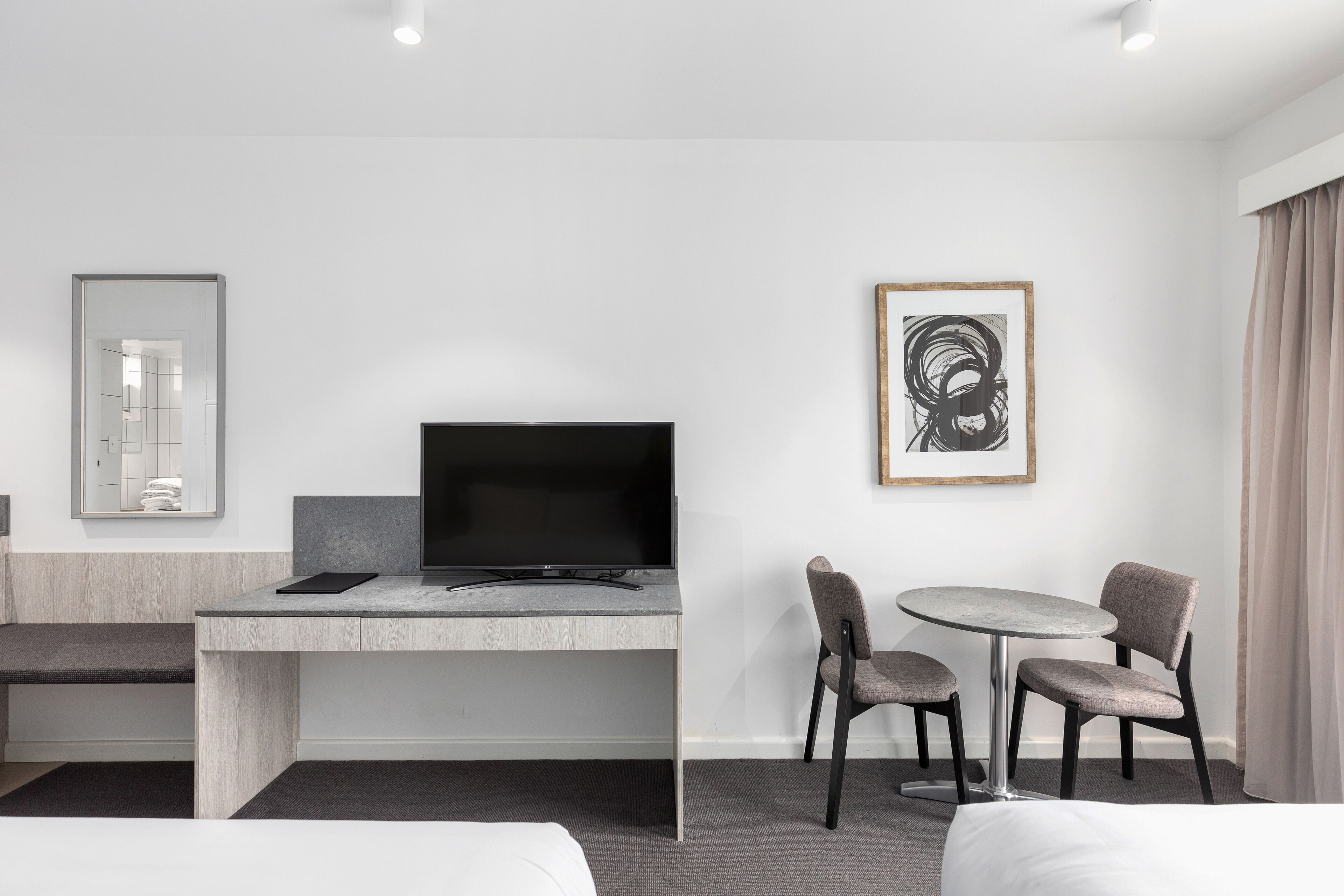 A modern hotel room with a bed, a desk with a flat-screen TV, a small round table with two chairs, and a framed abstract artwork on the wall.