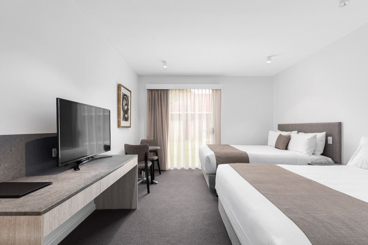A hotel room with a flat-screen TV, a desk with a chair, two beds with pillows, and a large window with curtains.
