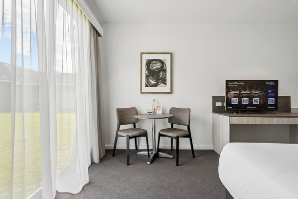 A hotel room with a bed, a TV, a table with two chairs, a framed artwork, and a window with sheer curtains.