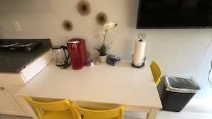 A kitchen with a white table, yellow chairs, a red coffee maker, a potted plant, and a TV on the wall.