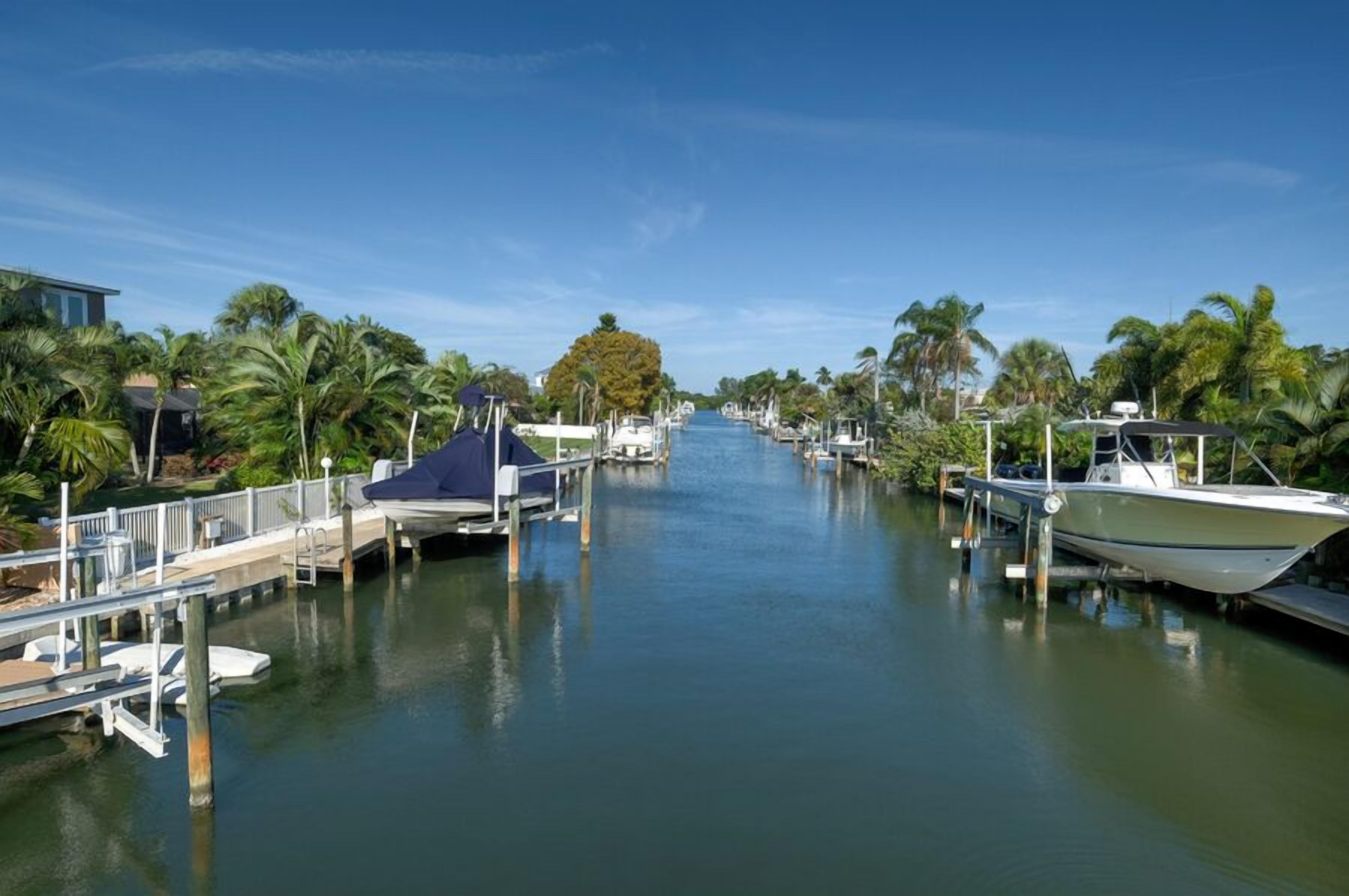 A marina with boats docked along a calm waterway, surrounded by palm trees and a clear sky.