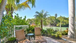 A patio with two wicker chairs and a small table, surrounded by lush greenery and a view of a lake. - Luxury waterfront home with a heated pool and spa! (Anna Maria)