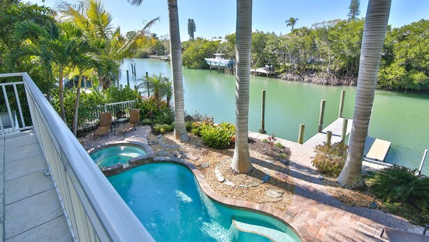 A swimming pool with a curved edge, surrounded by palm trees and a view of a calm body of water with a dock and boat. - Luxury waterfront home with a heated pool and spa! (Anna Maria)
