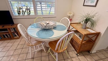 Un coin repas avec une table ronde, des chaises, une bibliothèque, une plante en pot et une fenêtre avec des stores.