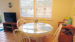 A dining area with a round glass table, white chairs, a television on a stand, and a window with blinds. - Delightful, comfortable villa just steps from the beach! (Holmes Beach)