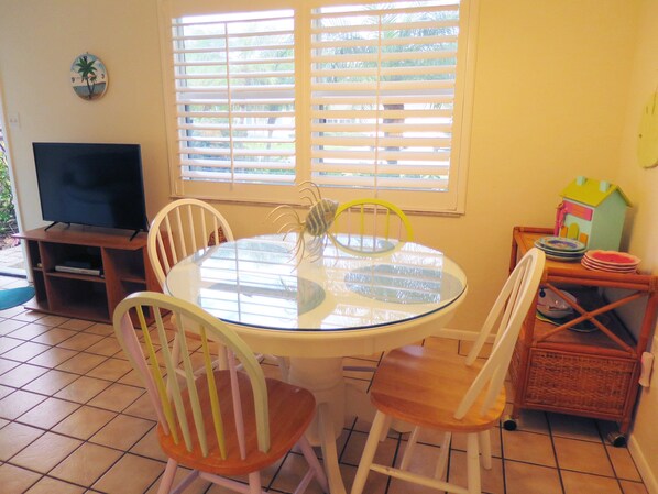 Un comedor con una mesa redonda de vidrio, sillas blancas, un televisor sobre un mueble bajo y una ventana con persianas.