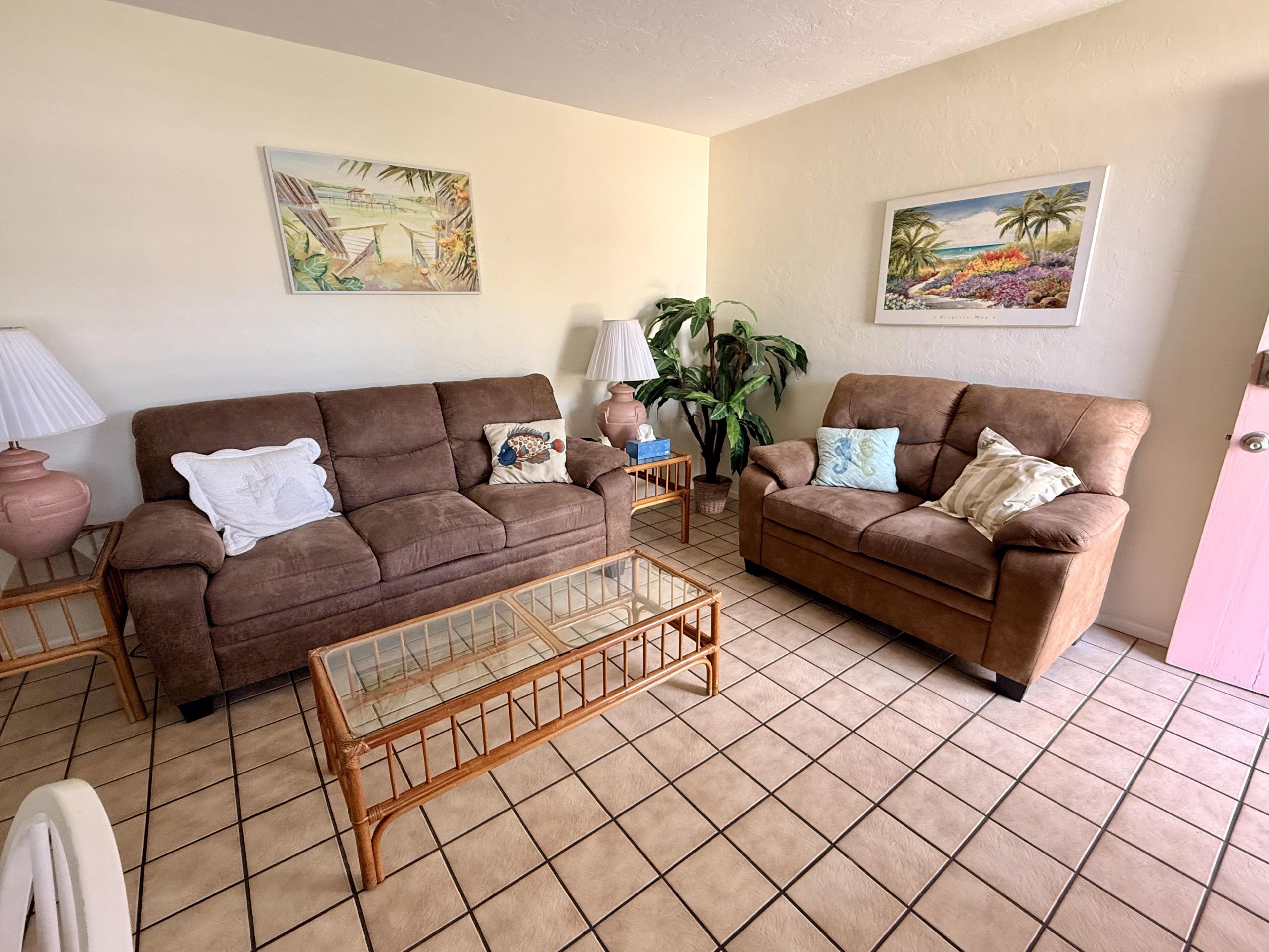 A living room with two brown sofas, a glass coffee table, and two wall-mounted paintings.