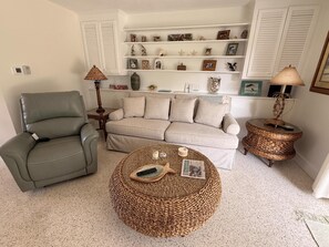 Una sala de estar con sofá, sillón, reposapiés redondo de mimbre y una estantería con adornos.
