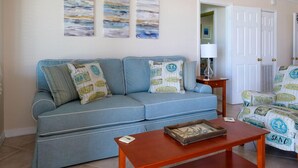 A living room with a blue sofa, a wooden coffee table, and a patterned armchair. There are two abstract paintings on the wall and a white door in the background.