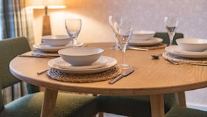 Una mesa redonda de madera para comer, dispuesta con cuencos blancos, copas de vino y cubiertos, rodeada de sillas tapizadas en verde.
