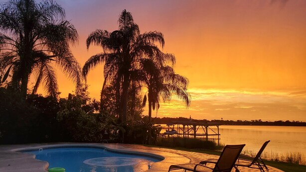A swimming pool with lounge chairs, surrounded by palm trees and a dock extending into a body of water, during sunset. - The Russell Lake House (Winter Haven)