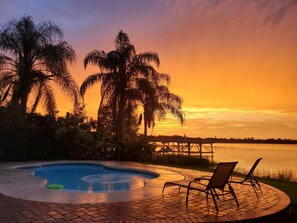 A swimming pool with lounge chairs, surrounded by palm trees and a dock extending into a body of water, during sunset. - The Russell Lake House (Winter Haven)