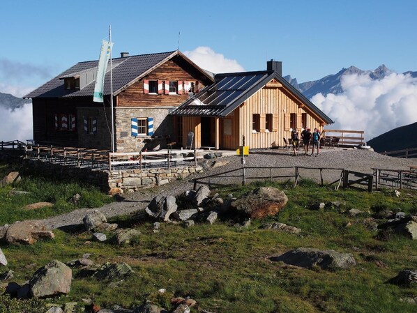 Situated on a rocky terrain, this mountain lodge offers a communal dining area with benches and tables outside, surrounded by breathtaking mountain views.