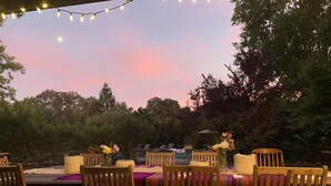 Un espace de restauration en plein air avec des chaises et des tables en bois, des guirlandes lumineuses et une vue sur des arbres et une piscine.