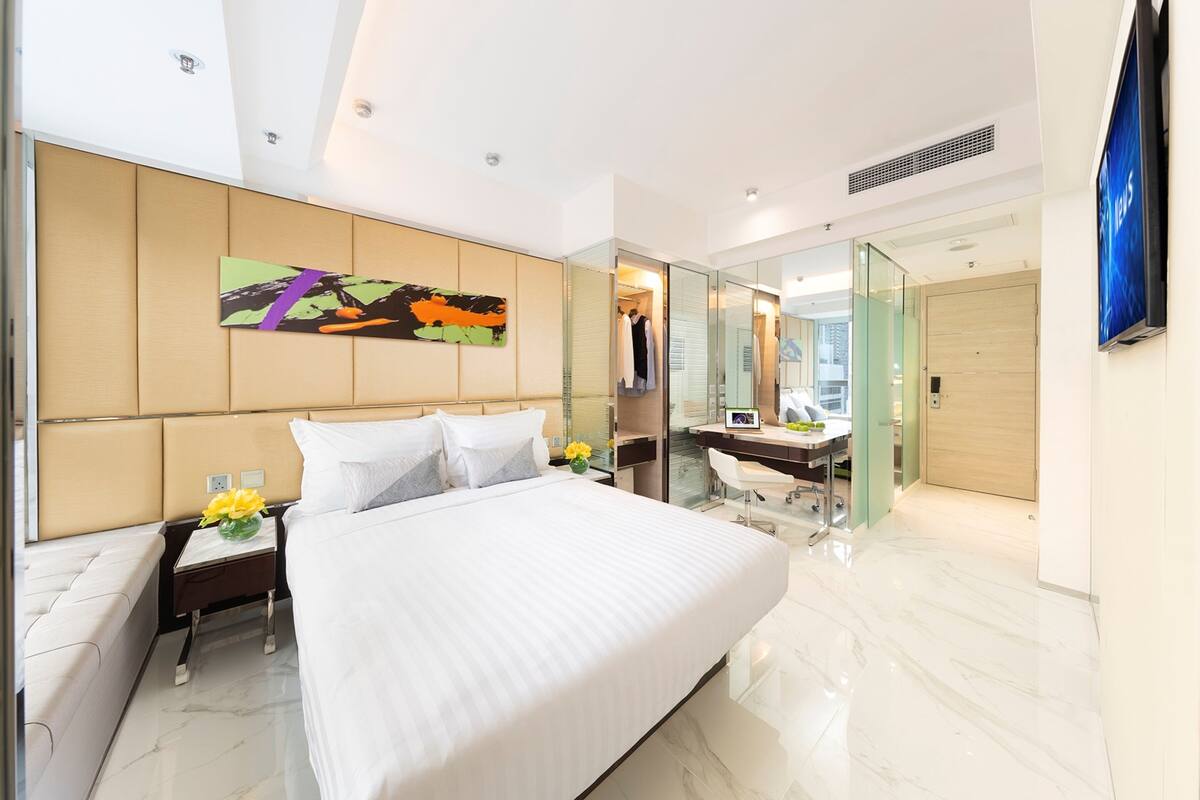 Una habitación de hotel moderna con una cama grande, una mesita de noche con un florero, un escritorio con una silla, un espejo grande y un televisor.