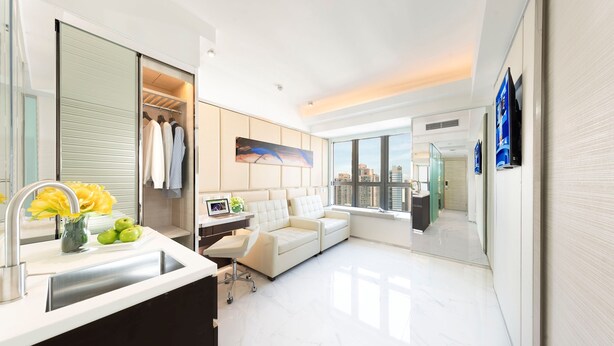 A modern hotel room with a white sofa, a desk with a chair, a large window, and a city view.