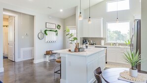 Una cocina moderna con armarios blancos, una isla central, iluminación colgante y vistas al exterior a través de un ventanal.