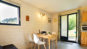 A cozy room with a wooden table, white chairs, a black sofa, a small wooden birdhouse, and a window with a view of greenery. - Le Bochate by Interhome (Les Bottières)
