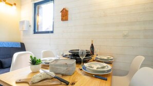 A dining area with a wooden table set for four, white chairs, a small potted plant, a cutting board with a knife, and a wine bottle. - Le Bochate by Interhome (Les Bottières)