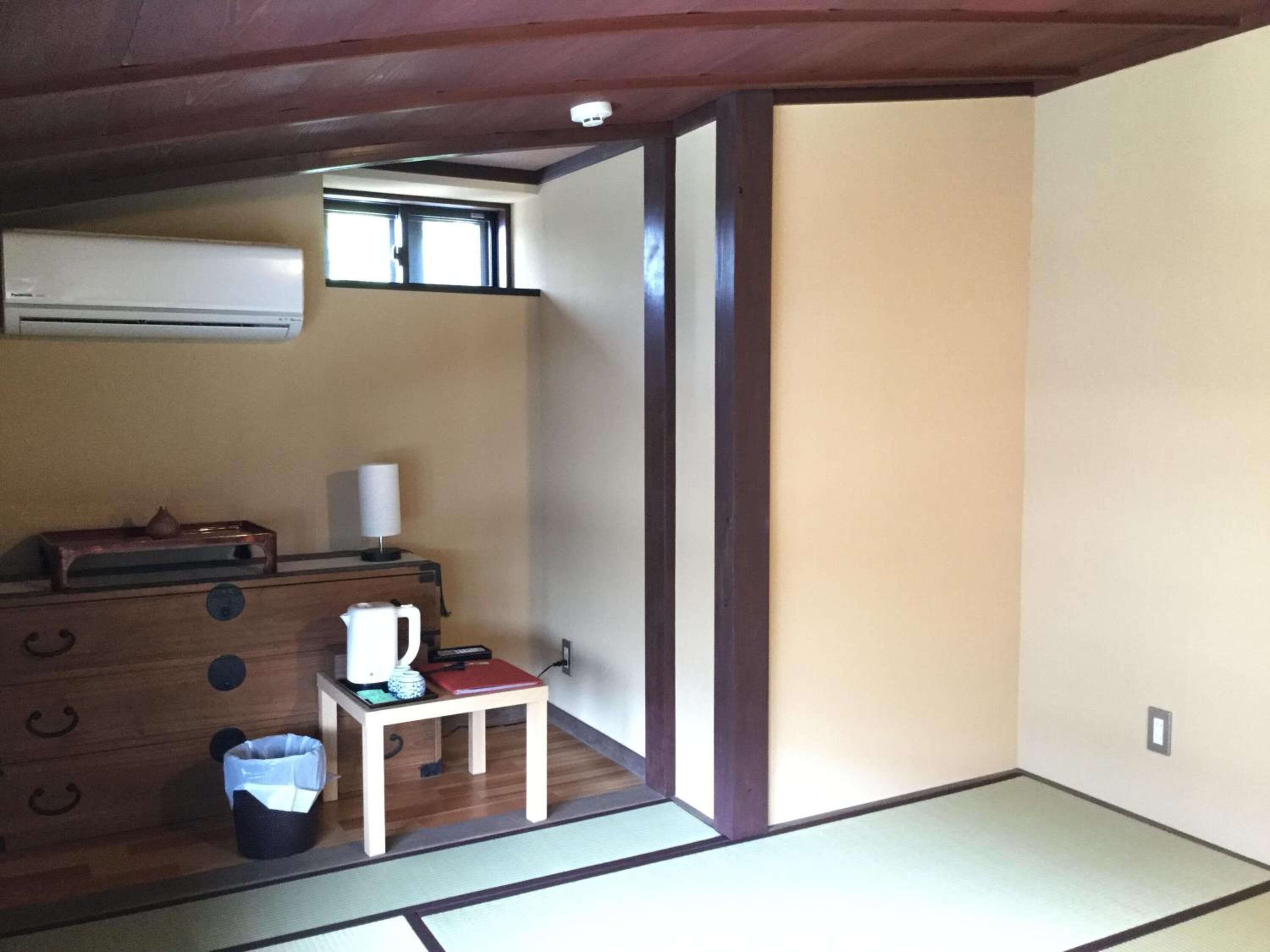 A small room with a wooden dresser, a small table with a kettle, a trash bin, and a window.