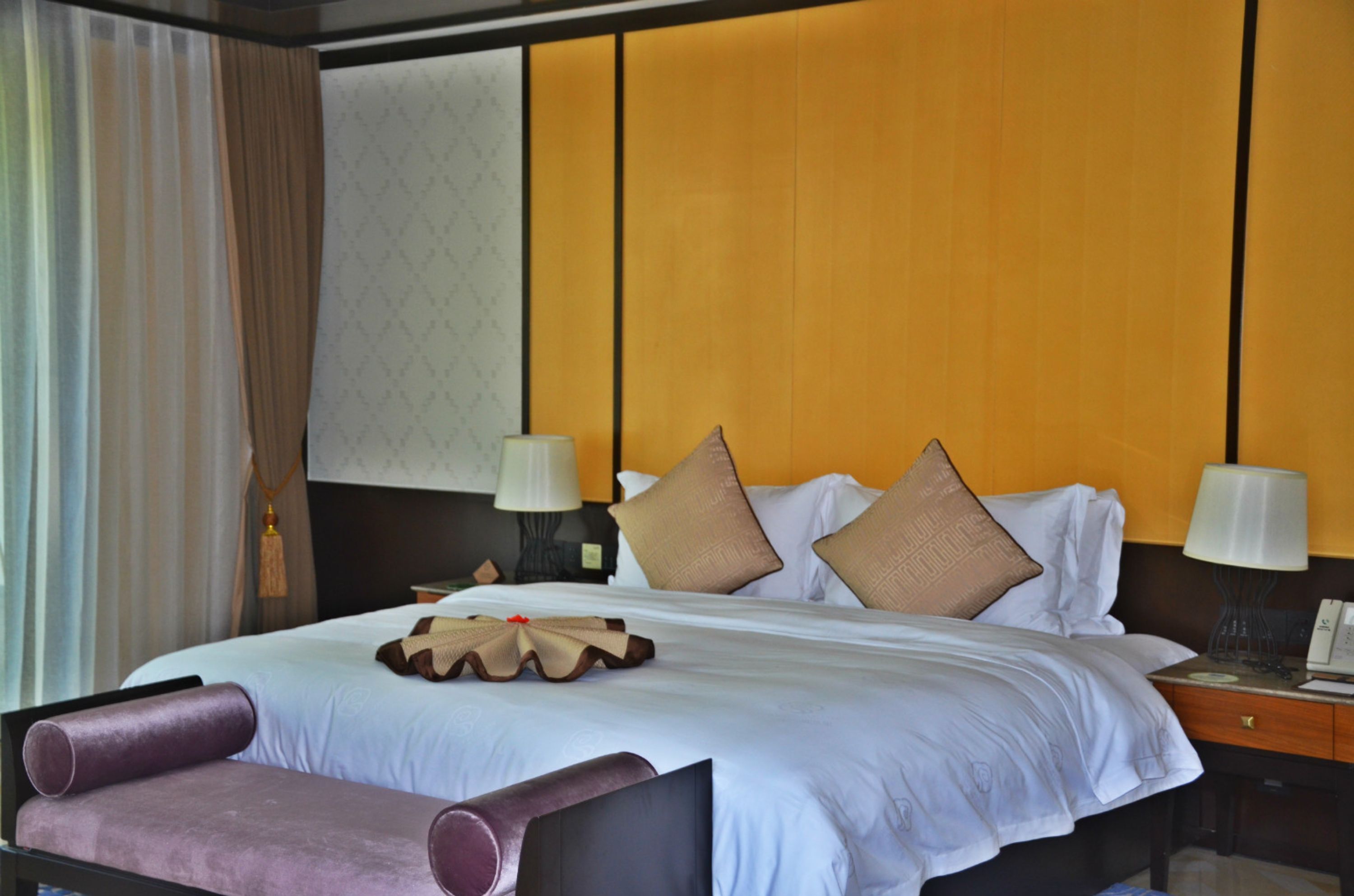 A hotel room with a large bed, two bedside tables with lamps, a bench at the foot of the bed, and a window with curtains.