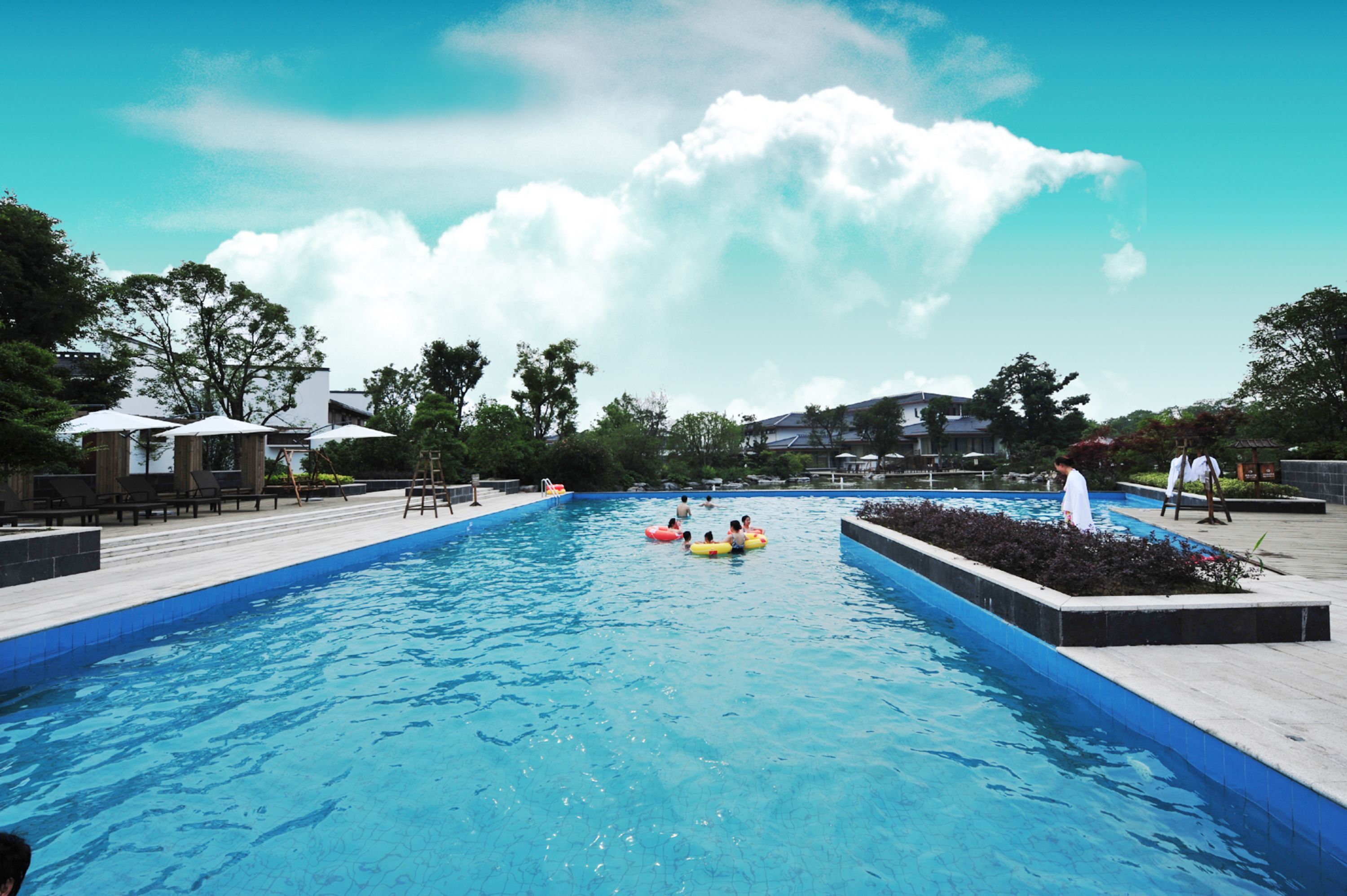 A swimming pool with lounge chairs and umbrellas, surrounded by trees and buildings.