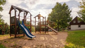Een speeltuin met een glijbaan en klimconstructies, omringd door zand en gras, met bomen en een huis op de achtergrond.