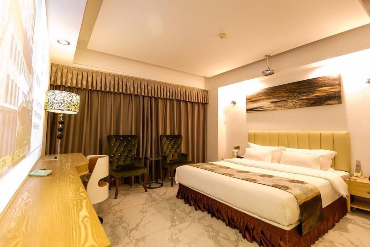 A hotel room with a bed, a desk, two chairs, a lamp, and a painting on the wall.