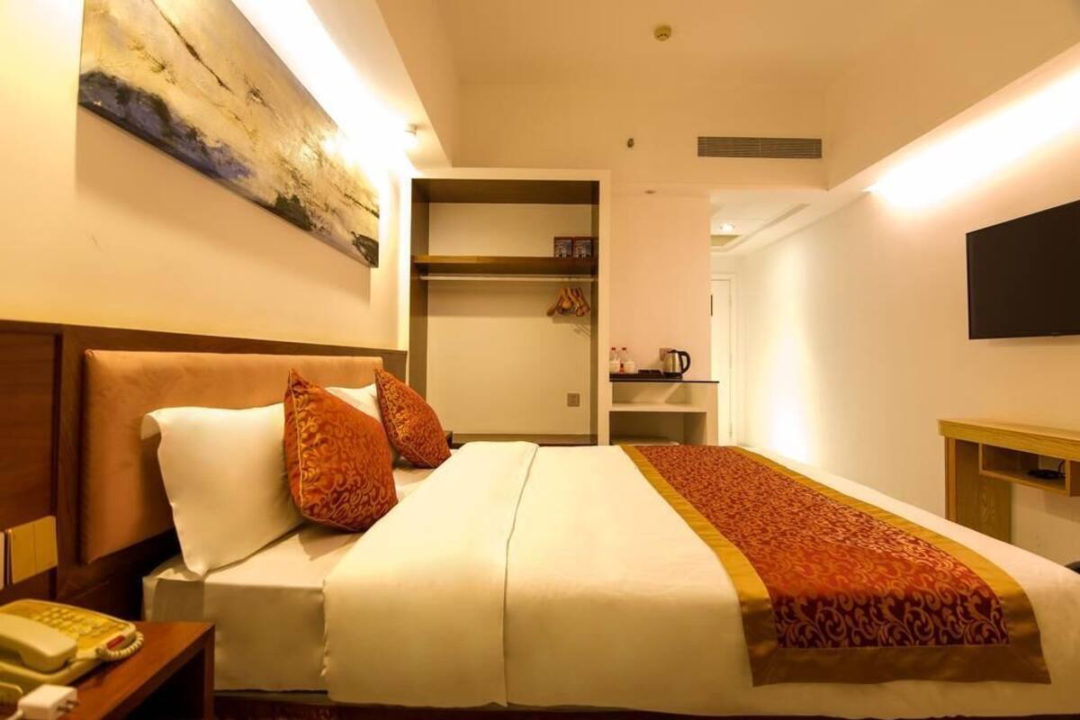 A hotel room with a bed, a bedside table with a telephone, a painting on the wall, a television mounted on the wall, and a shelf with items.