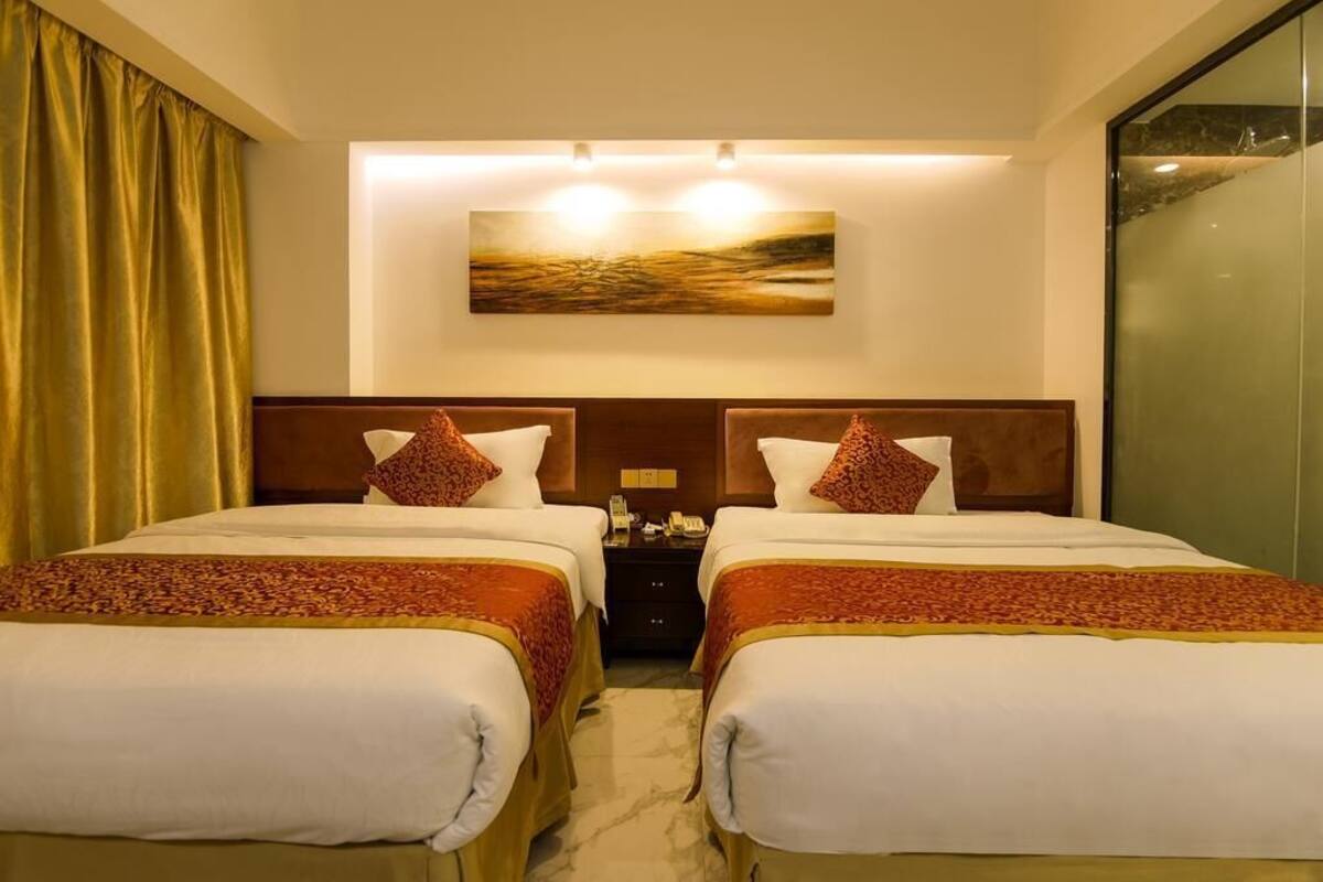 A hotel room with two beds, a nightstand, a painting above the beds, and a view through a glass door.