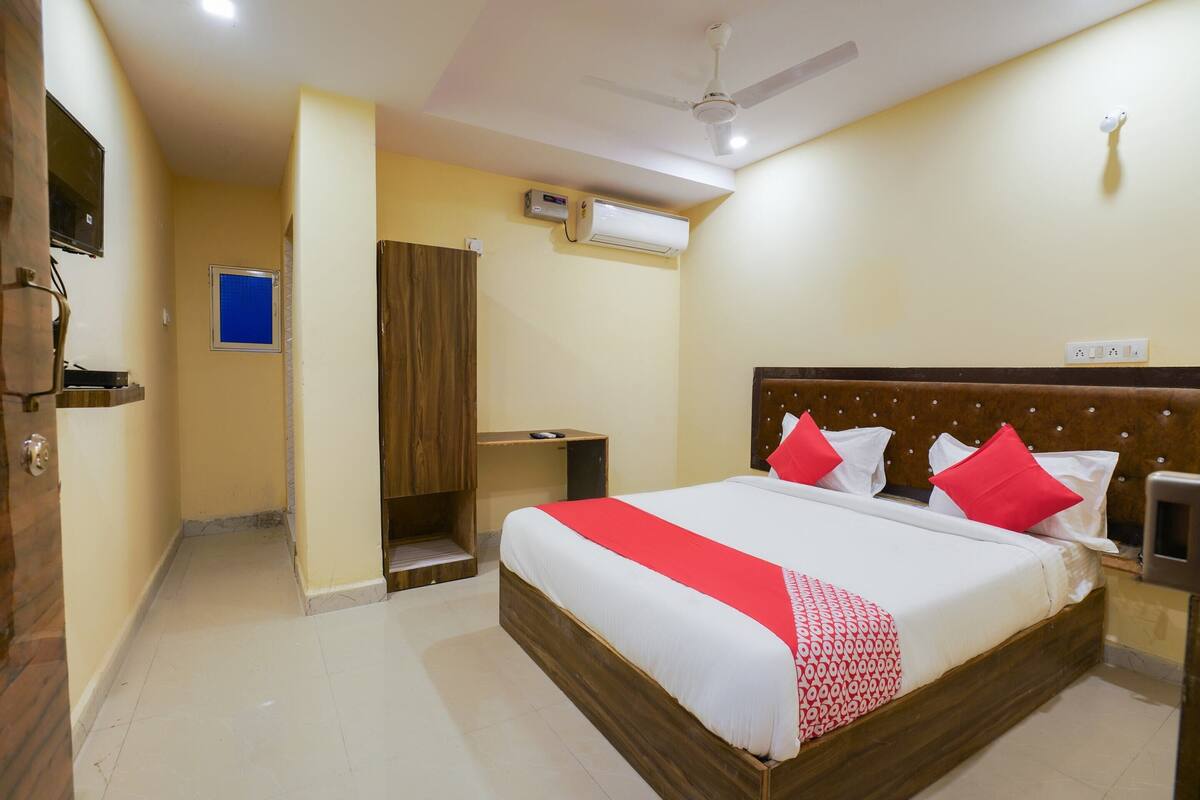 A hotel room with a large bed, a wooden headboard, a ceiling fan, an air conditioning unit, and a small desk with a chair.