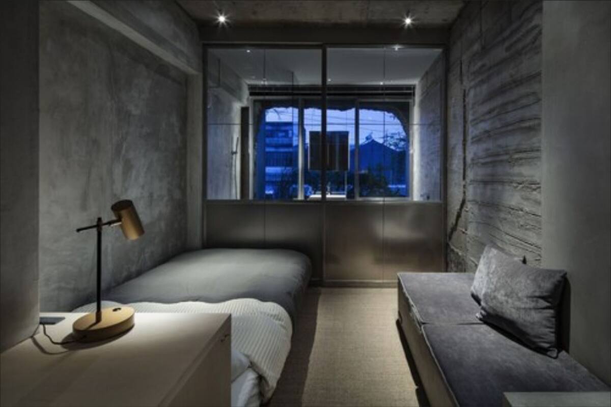 A bedroom with a bed, a bedside table, a lamp, a sofa, and a window with a view of buildings.