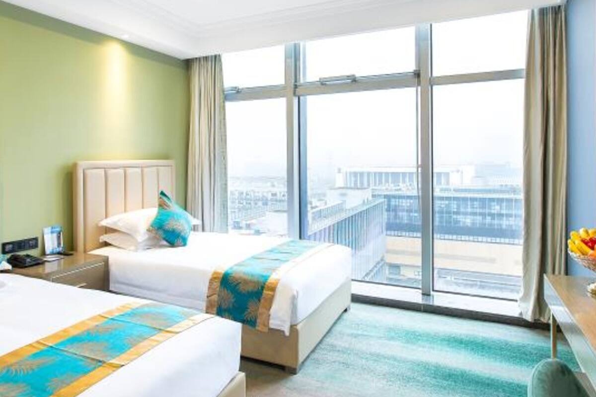 A hotel room with two beds, a desk, and a large window offering a city view.