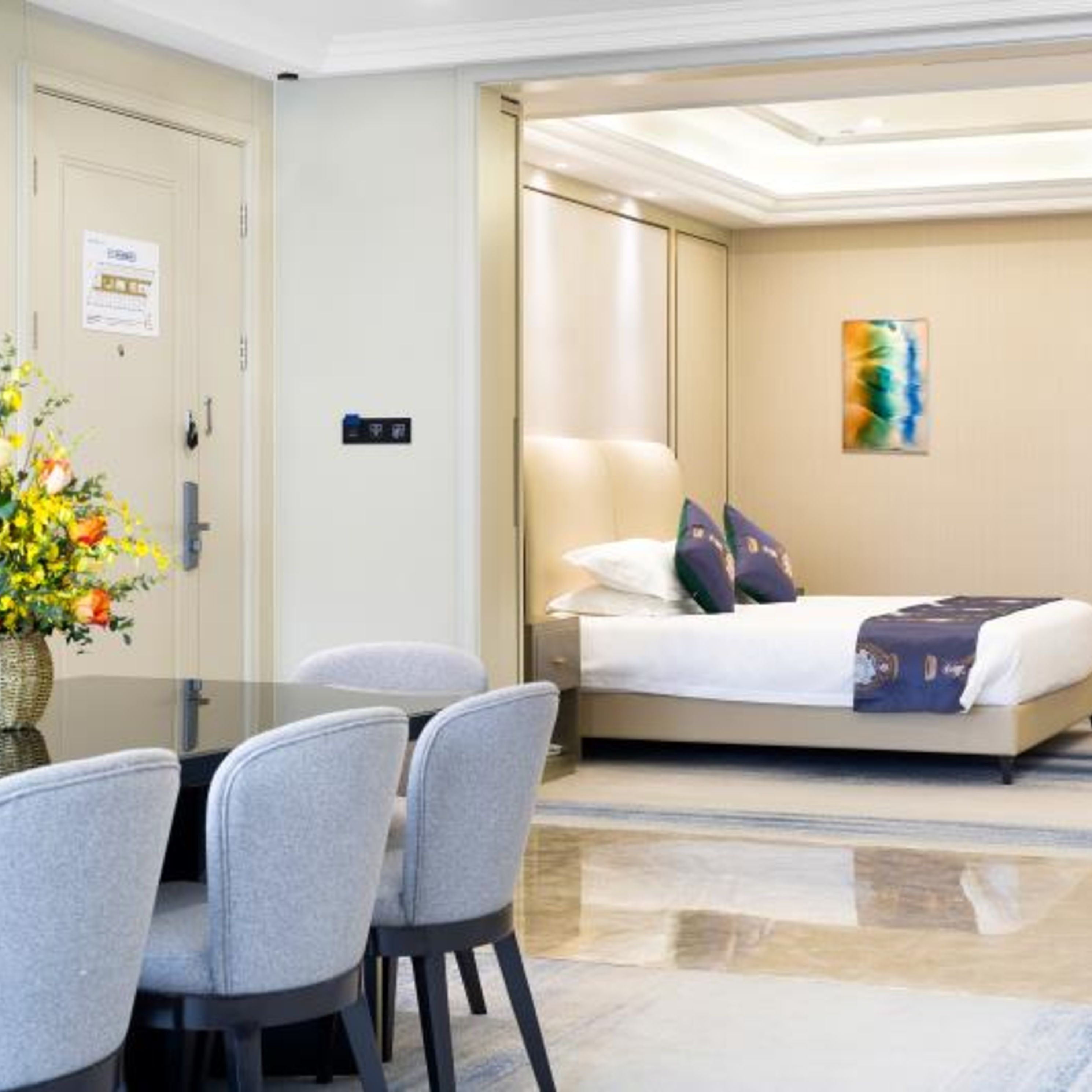 A hotel room with a bed, a desk, and chairs. There is a vase with flowers on the desk.