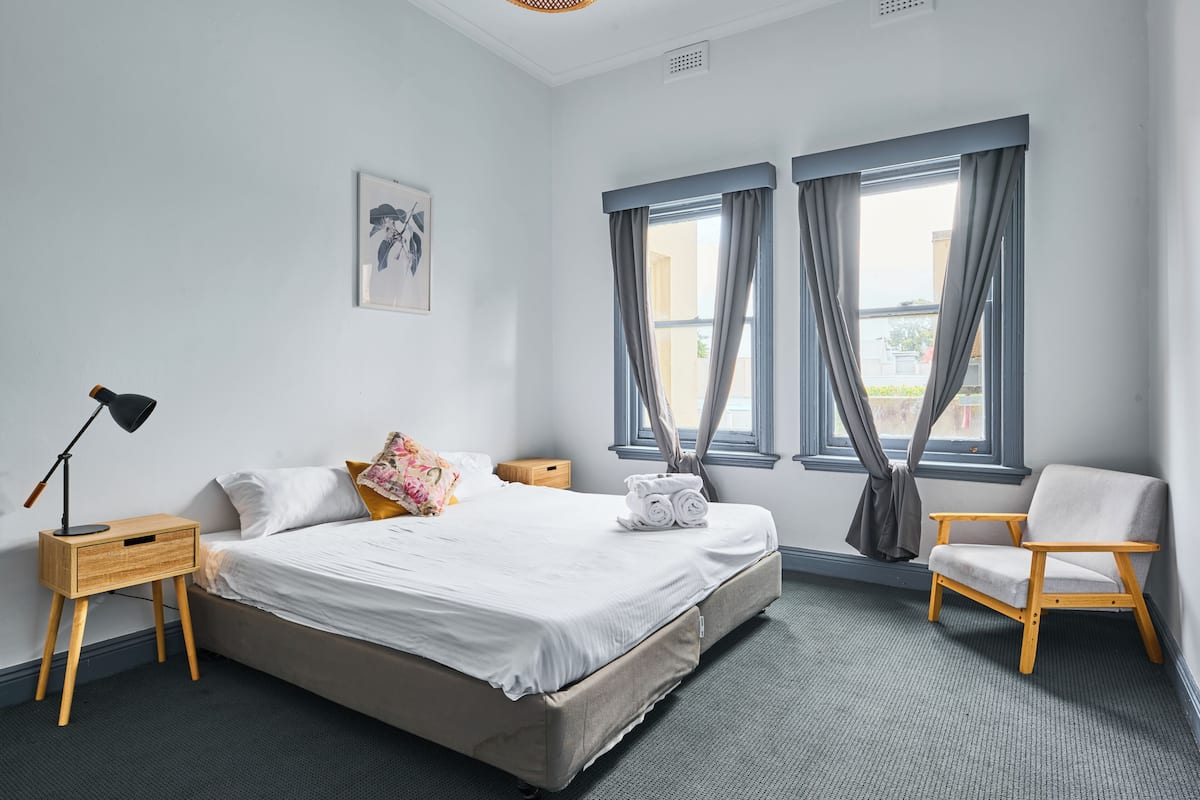 Habitación de hotel ordenada con cama, mesita de noche, silla y dos ventanas con cortinas.