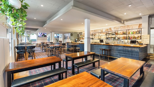 A restaurant with wooden tables and benches, a bar area with shelves of bottles, and a television screen in the background. - College Lawn Hotel - Hostel (Prahran)